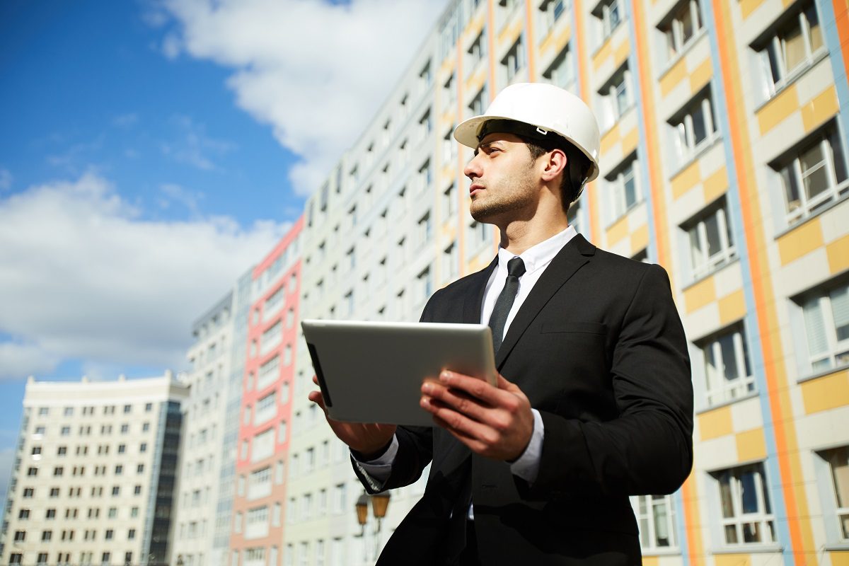 A land developer with a tablet and hard hat stands in front of a new apartment building.