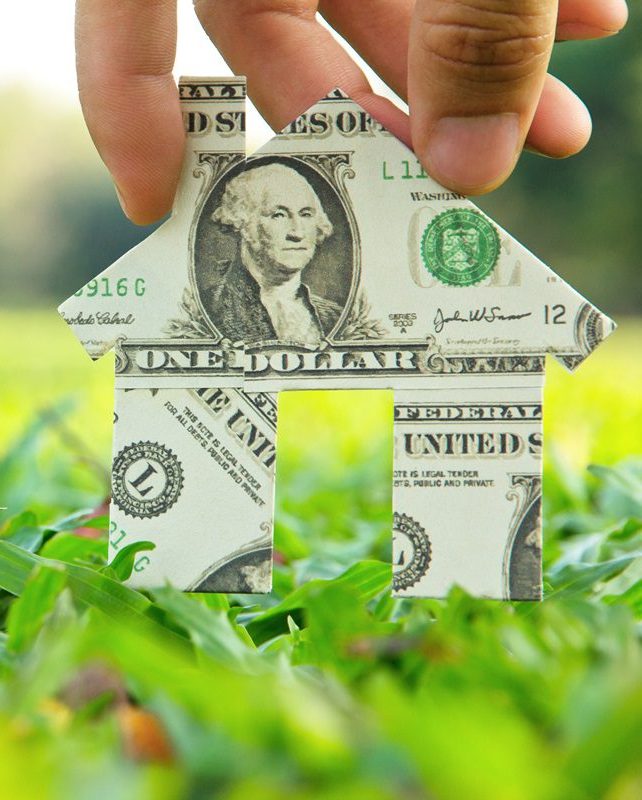 A person holding a dollar folded into a house shape on a lawn