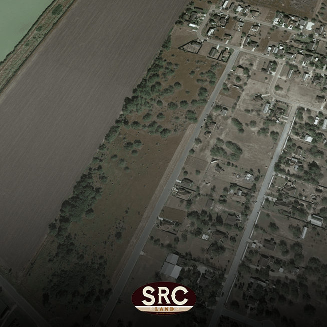 Aerial view of a rural area: a wide plowed field or shoreline on the left, a tree line in the middle, and a residential street grid on the right with houses; SRC Land logo at the bottom.