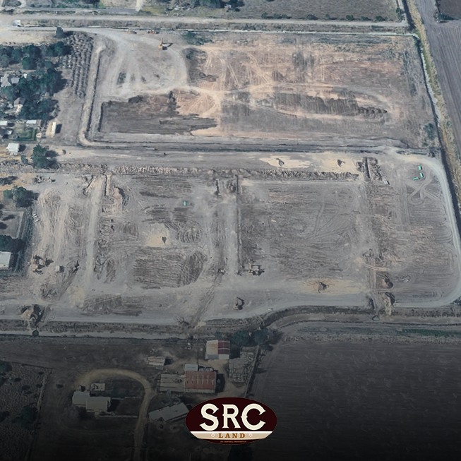 Aerial view of a large construction site with multiple cleared lots, dirt roads, and several excavators working; residential buildings visible at bottom left and an SRC Land logo lower center.